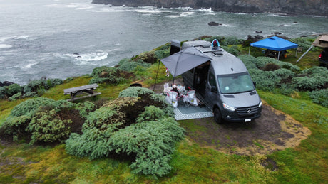 RV rental van parked on a coastal cliff, awning extended. People enjoy the ocean view in this scenic outdoor camp setup.