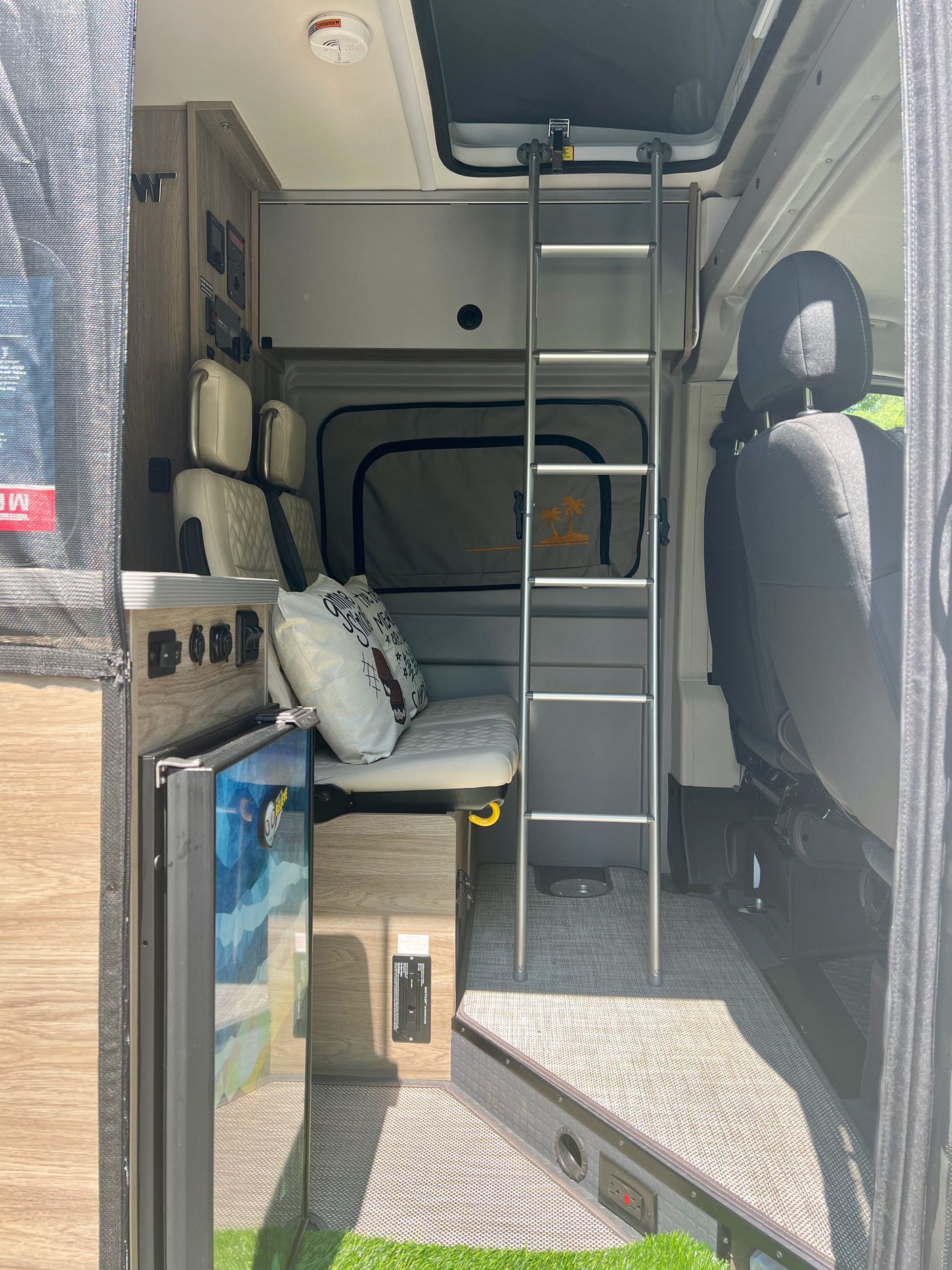 View inside a modern RV rental featuring a bunk bed, bench seating, compact fridge, and control panel for road trip comfort.
