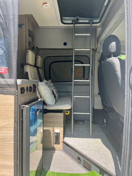 View inside a modern RV rental featuring a bunk bed, bench seating, compact fridge, and control panel for road trip comfort.