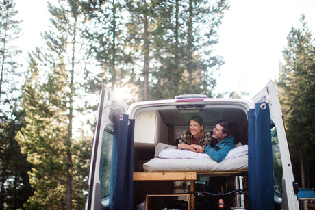 Happy couple enjoying their RV rental adventure, relaxing in a custom camper van parked in a sunny forest.