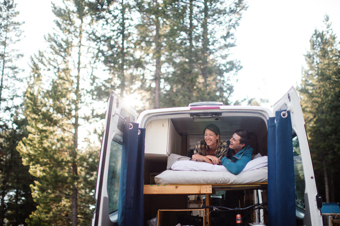Happy couple enjoying their RV rental adventure, relaxing in a custom camper van parked in a sunny forest.