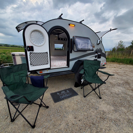 This RV rental teardrop camper is set up for a relaxing stay, with open door revealing the bed, chairs, and a welcome mat outside.