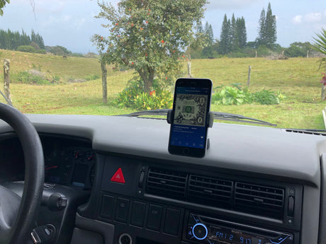 Inside a classic RV rental, a phone mounted on the dash plays music. Outside, a picturesque rural landscape with green fields and grazing animals.