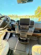 Inside a modern RV rental: driver's view of the dashboard, steering wheel, and scenic waterfront with buildings and a palm tree.