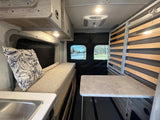This rv rental interior has a cozy bench, sink, table, and slatted bed frame, with bright overhead lights and outdoor views.