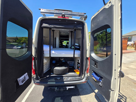 RV rental: Open rear doors reveal a versatile camper van interior with a bed, under-bed storage, and utility access.