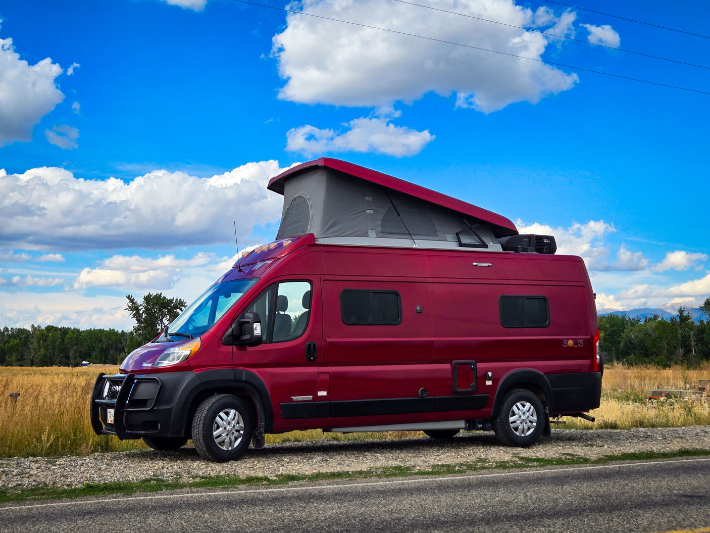A red Winnebago Solis camper van, available for RV rental, parked roadside with a pop-top roof extended, under a blue sky.