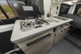 Inside this RV rental: a compact kitchen with a two-burner stove, sink, and counter, next to a cozy bed.