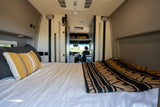 Interior of an RV rental, showing a cozy bed with pillows and a blanket, looking forward towards the driver's seat.