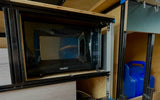 RV rental: Compact microwave integrated into a custom camper van kitchen with blue water jugs for mobile living.