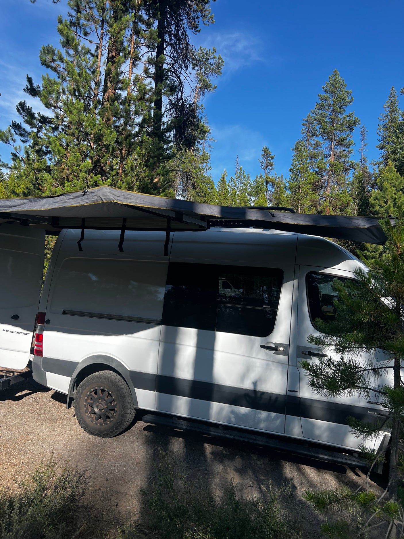 White camper van with extended awning parked in a forest campsite under a blue sky – your next RV rental adventure.