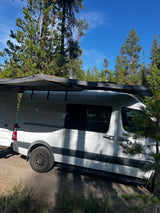 White camper van with extended awning parked in a forest campsite under a blue sky – your next RV rental adventure.