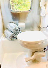 An RV rental's compact bathroom showing a toilet, neatly rolled towels, and a window looking out to green trees.
