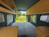 Cozy RV rental interior features a comfortable bed, wooden storage, and windows with curtains overlooking a serene outdoor view.