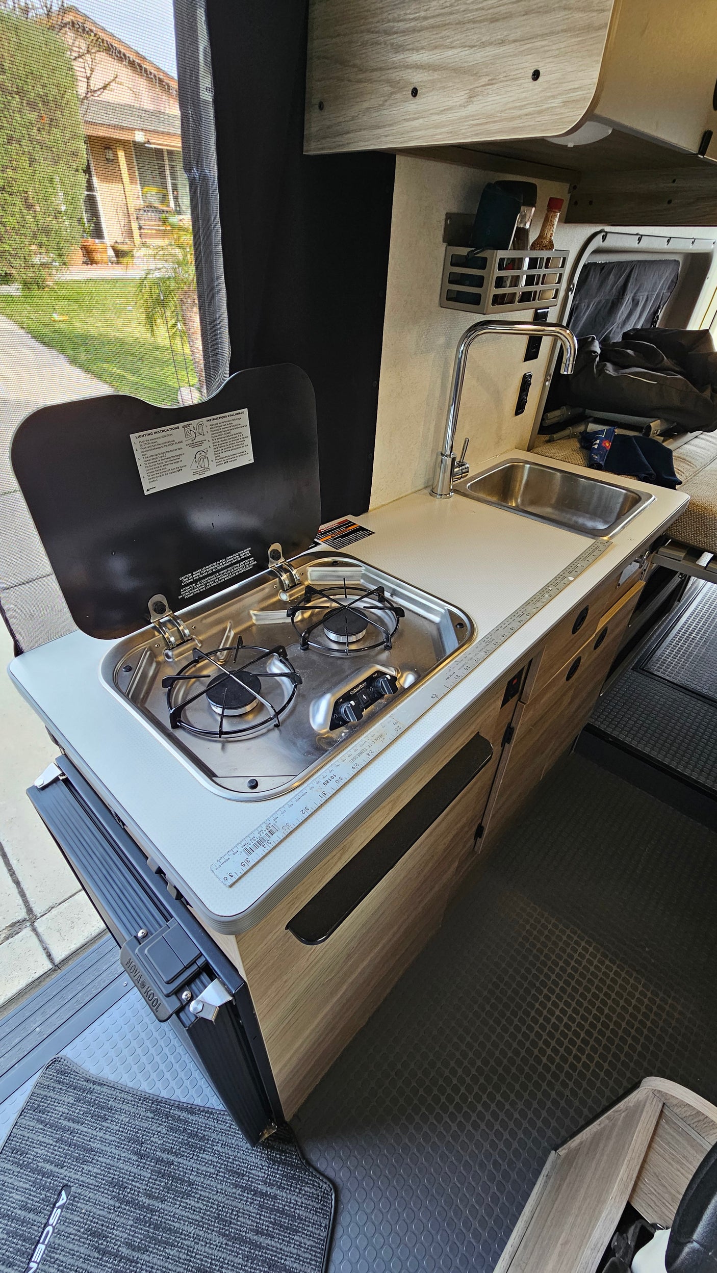 An RV rental's compact kitchen, featuring a two-burner stove, sink, and faucet, offers a convenient setup with an outdoor view.