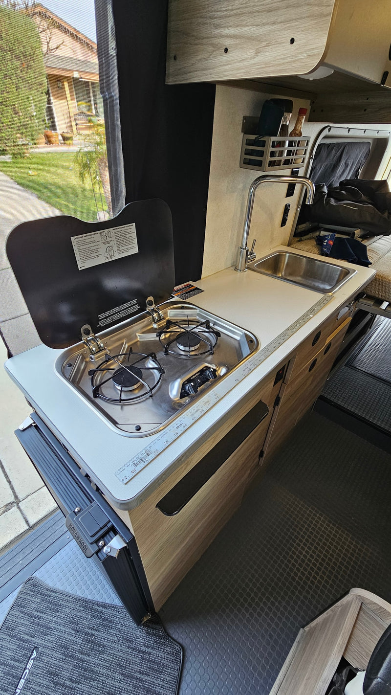 An RV rental's compact kitchen, featuring a two-burner stove, sink, and faucet, offers a convenient setup with an outdoor view. — photo 7