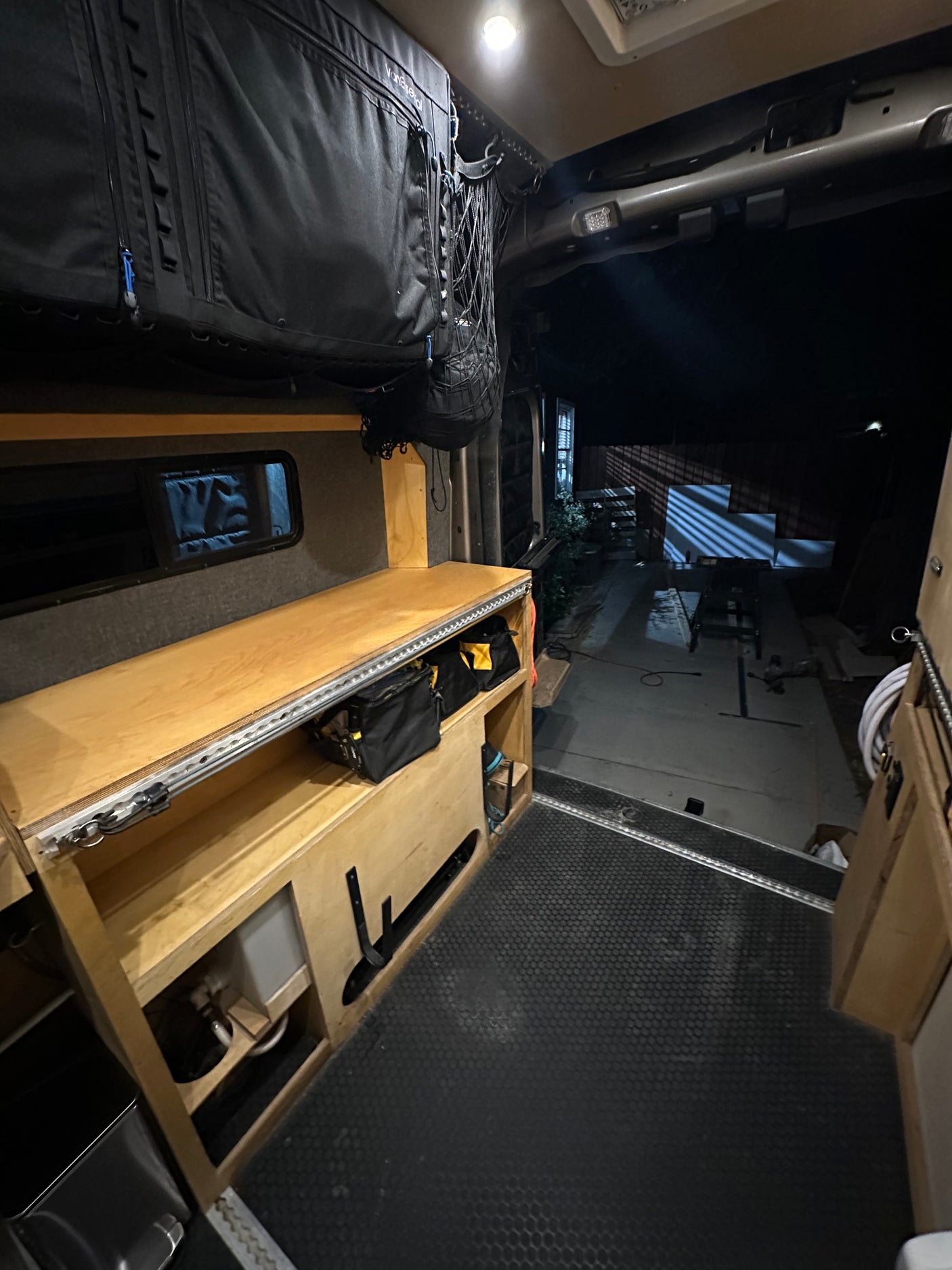 RV rental interior: an overhead storage bag, modular wooden shelving with tools, and an open rear hatch revealing a dark outdoor area.