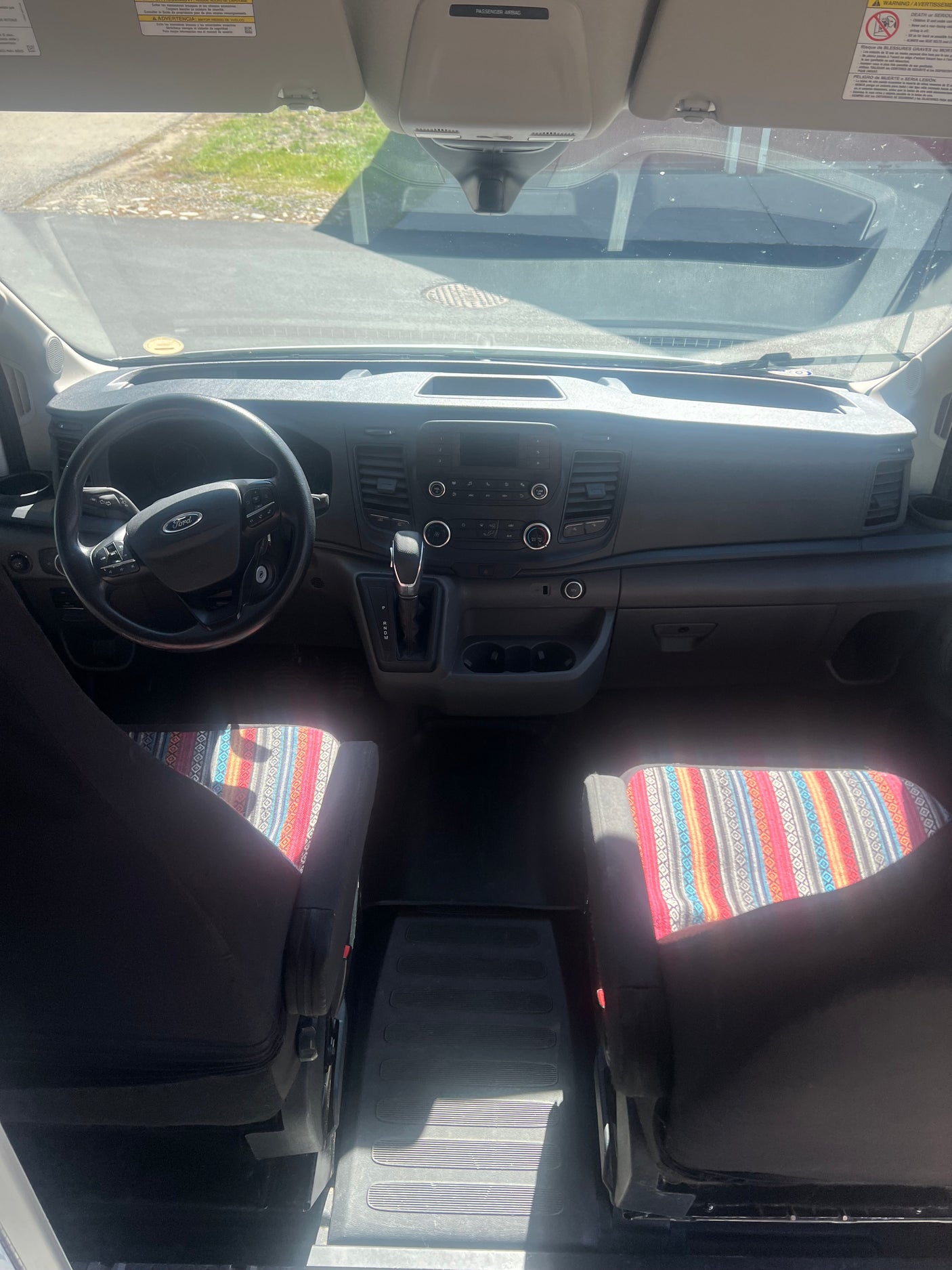 Front cabin of an rv rental van with Ford dashboard, steering wheel, gear shifter, and vibrant patterned seat covers.