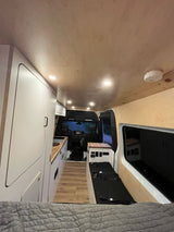 Modern RV rental van conversion featuring bright white cabinetry, wood counters, recessed lighting, and a cozy bed.