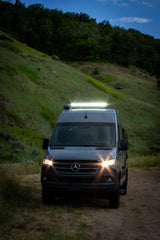This dark grey Mercedes Sprinter camper van, a bright RV rental, illuminates a dirt path at dusk in a hilly landscape.