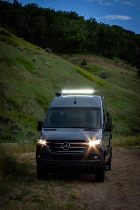 This dark grey Mercedes Sprinter camper van, a bright RV rental, illuminates a dirt path at dusk in a hilly landscape.