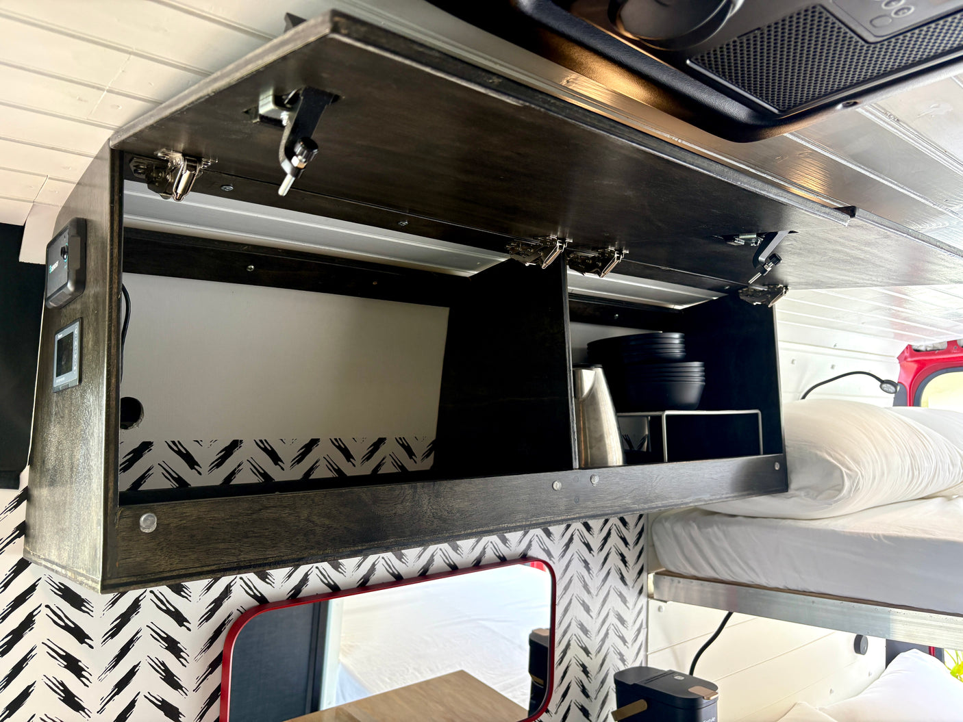 This RV rental overhead cabinet features secure latches, black dishes, storage, controls, and a cozy bed with a chevron accent wall.