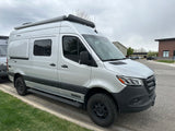 A silver Winnebago Revel camper van, an ideal rv rental, with rugged tires and roof awning, parked on a street.