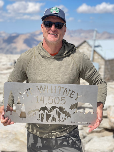 Hiker celebrates reaching Mt. Whitney's 14,505 ft summit, holding a commemorative sign. Ideal destination for an RV rental!