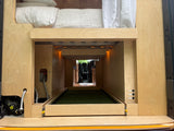 Rear of a custom RV rental van: raised bed, large storage with pull-out turf tray, water pump, and electrical controls.