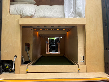 Rear of a custom RV rental van: raised bed, large storage with pull-out turf tray, water pump, and electrical controls.