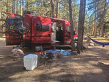 Red RV rental van with bikes parked in a sunlit pine forest campsite. A blue hammock hangs nearby, with a small dog and cooler visible.