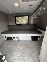 Cozy RV rental interior with a gray sofa bed, large window, ceiling AC unit, and light wood-tone flooring and cabinetry.