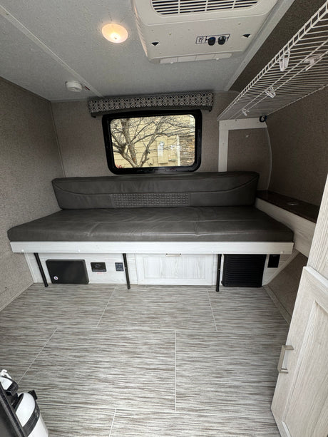 Cozy RV rental interior with a gray sofa bed, large window, ceiling AC unit, and light wood-tone flooring and cabinetry.