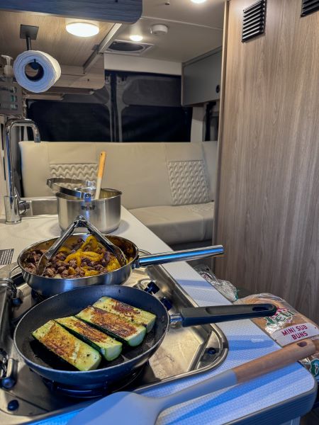 Inside a modern RV rental, a compact kitchen features pans of grilling zucchini and a meat skillet.