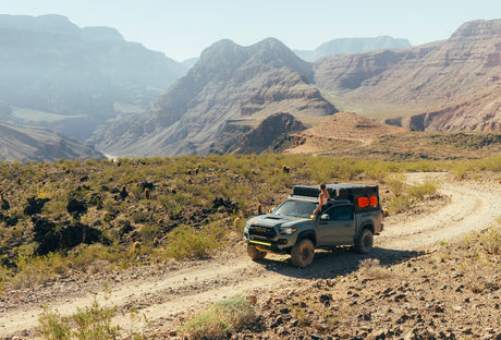 This customized overlanding truck, ideal for an rv rental adventure, navigates a desert trail with a person admiring canyon views.