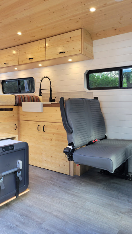 RV rental: Bright wood interior with kitchen, a cozy bed, gray folding seat, and cooler on grey wood floor.
