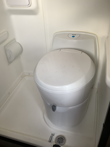 Inside an RV rental's compact, all-white bathroom: A Thetford portable toilet sits above a shower pan with drain.