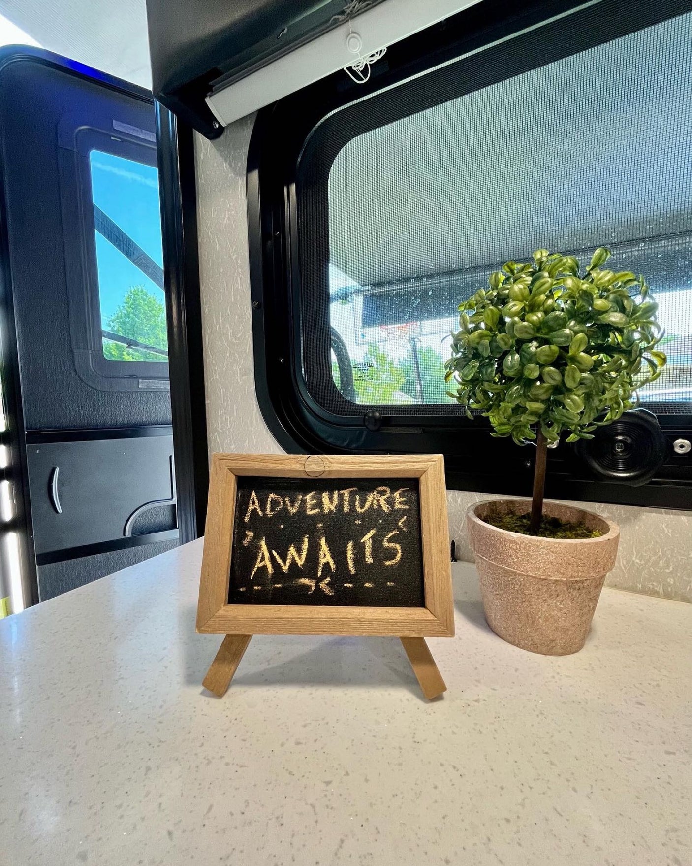 Cozy RV rental interior featuring a counter with an 'ADVENTURE AWAITS' sign, a small plant, and an open door to nature.