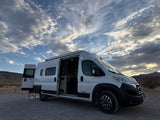 White Winnebago Solis RV rental with side and rear doors open, parked on gravel in a desert mountain landscape.