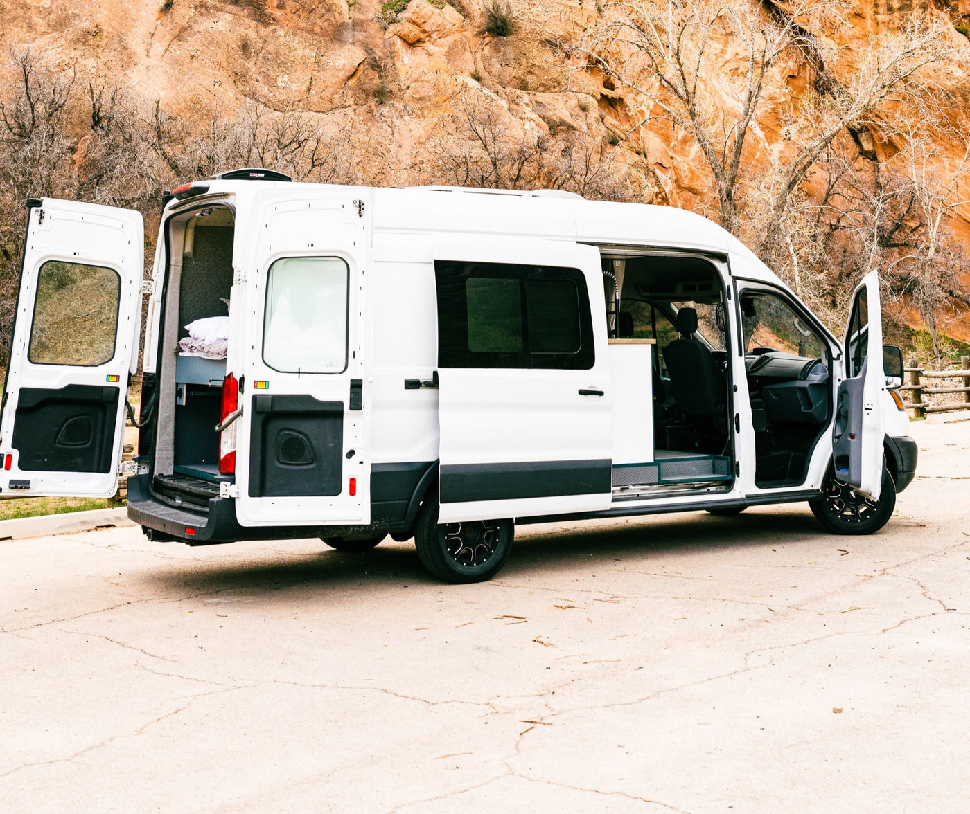 White RV rental van with all its doors open, showing the converted interior, parked against orange rock cliffs and trees. — photo 2