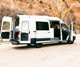 White RV rental van with all its doors open, showing the converted interior, parked against orange rock cliffs and trees.