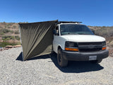 This white RV rental camper van with an olive green awning is set up by a pond for adventure in a sunny, gravelly landscape.