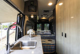 Inside a modern RV rental: compact kitchen with sink, comfy bed, and views of nature through the open door.