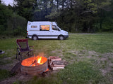 Cozy night camping with an RV rental: a white camper van, crackling campfire, and a chair on green grass near trees.