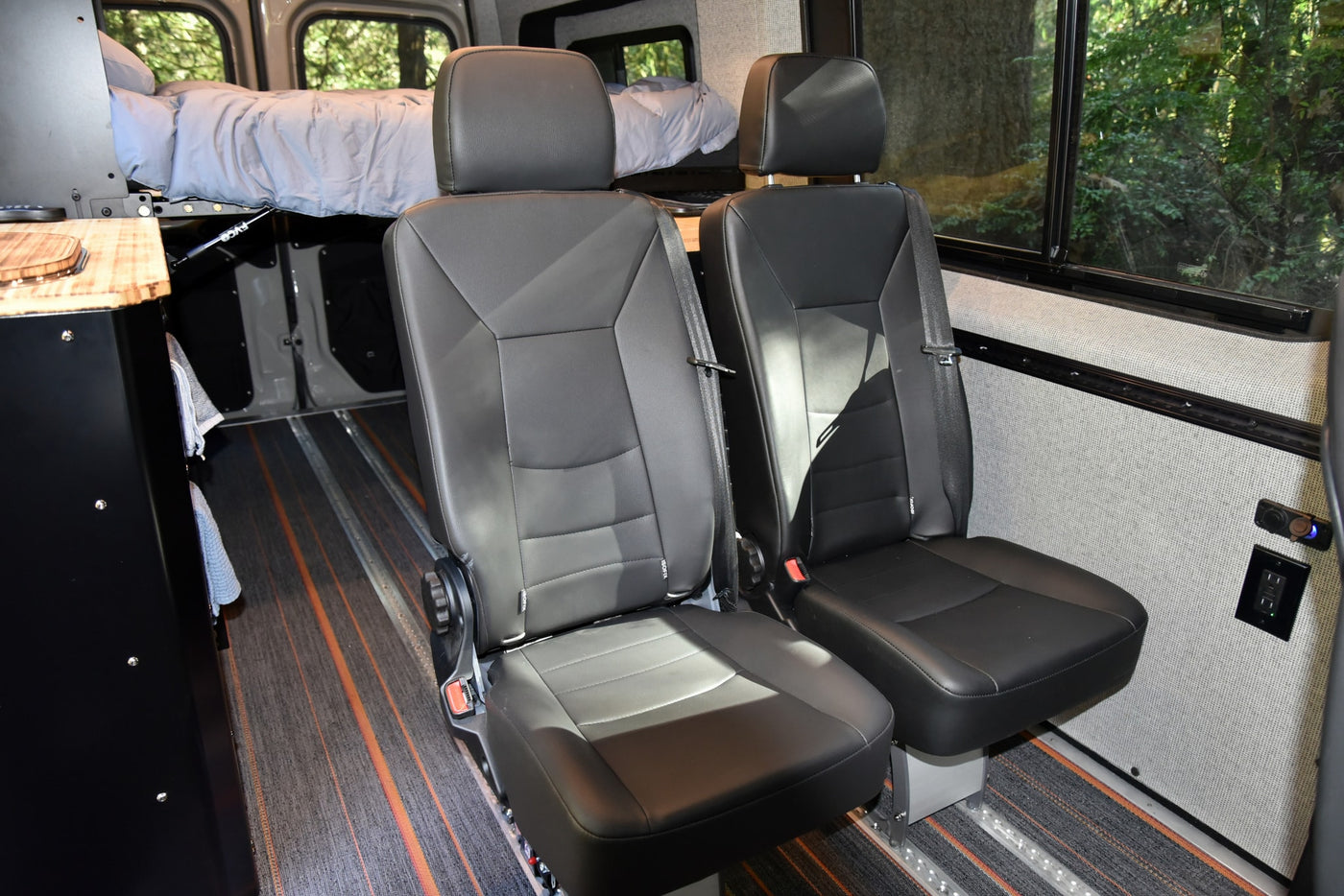 Modern RV rental camper van interior with two black passenger seats, a raised bed, and power outlets on the wall. — photo 12