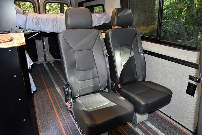 Modern RV rental camper van interior with two black passenger seats, a raised bed, and power outlets on the wall. — photo 12