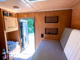 Cozy RV rental interior with wooden walls, open door to nature, comfy bed, and storage. Ideal for travel.