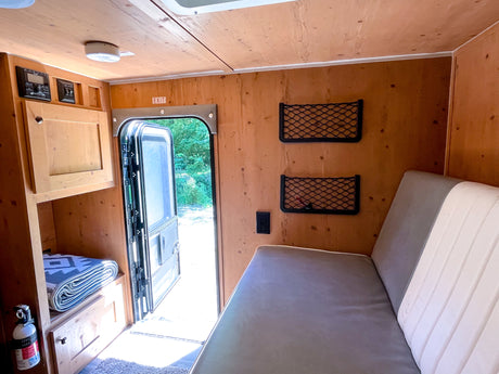 Cozy RV rental interior with wooden walls, open door to nature, comfy bed, and storage. Ideal for travel.