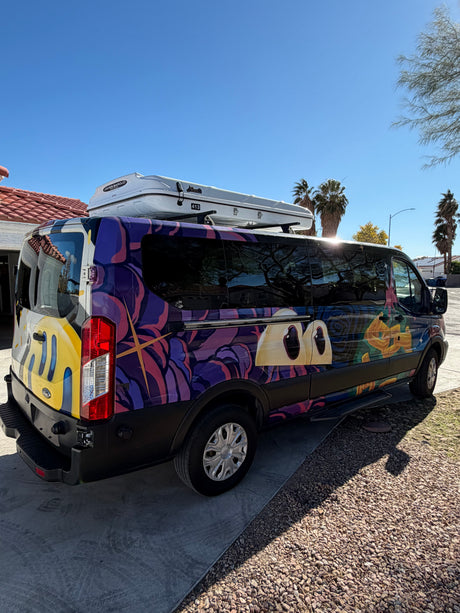 A colorful custom-wrapped RV rental van with abstract art, large eyes, and a rooftop cargo box, parked on a sunny day.