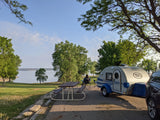 Lakeside campsite featuring a T@G teardrop RV rental, picnic table, and person relaxing by the water.
